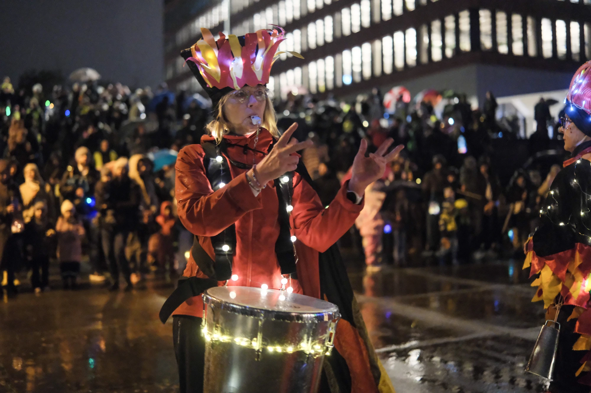 Person in rotem Kostüm mit leuchtender Trommel und bunter Kopfbedeckung bei Nachtveranstaltung mit Publikum im Hintergrund