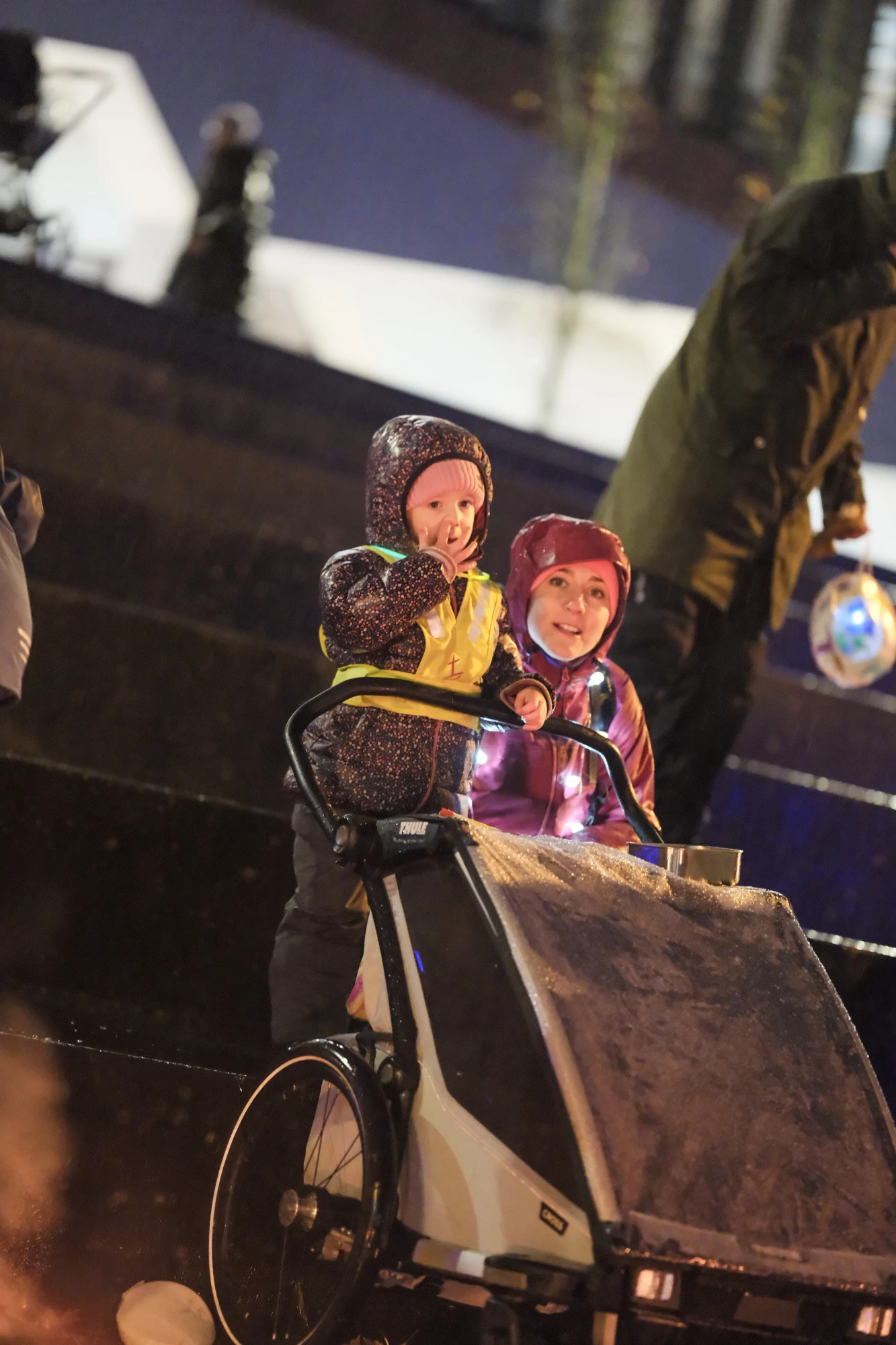 Zwei Kinder in wetterfester Kleidung sitzen in einem Kinderwagen, eine Person im Hintergrund hält eine Laterne.