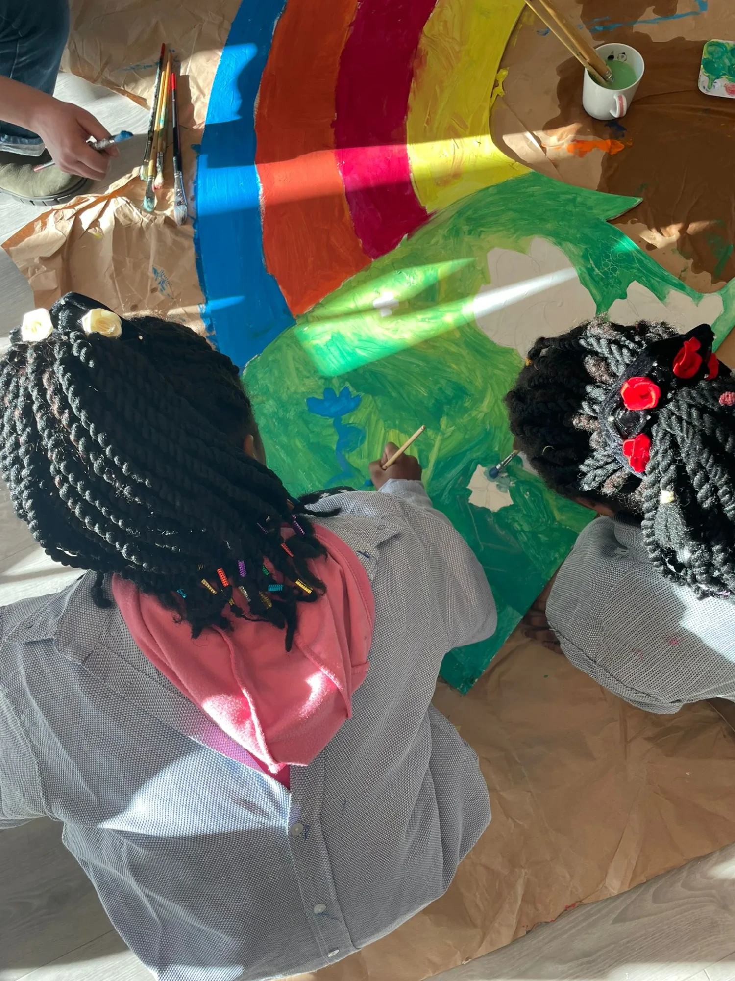 Zwei Kinder hocken auf dem boden und malen an einem Bild. Man sieht sie nur von hinten. DAs Bild Bild, das sie malen ist grün und darüber ist ein Regenbogen zu sehen.