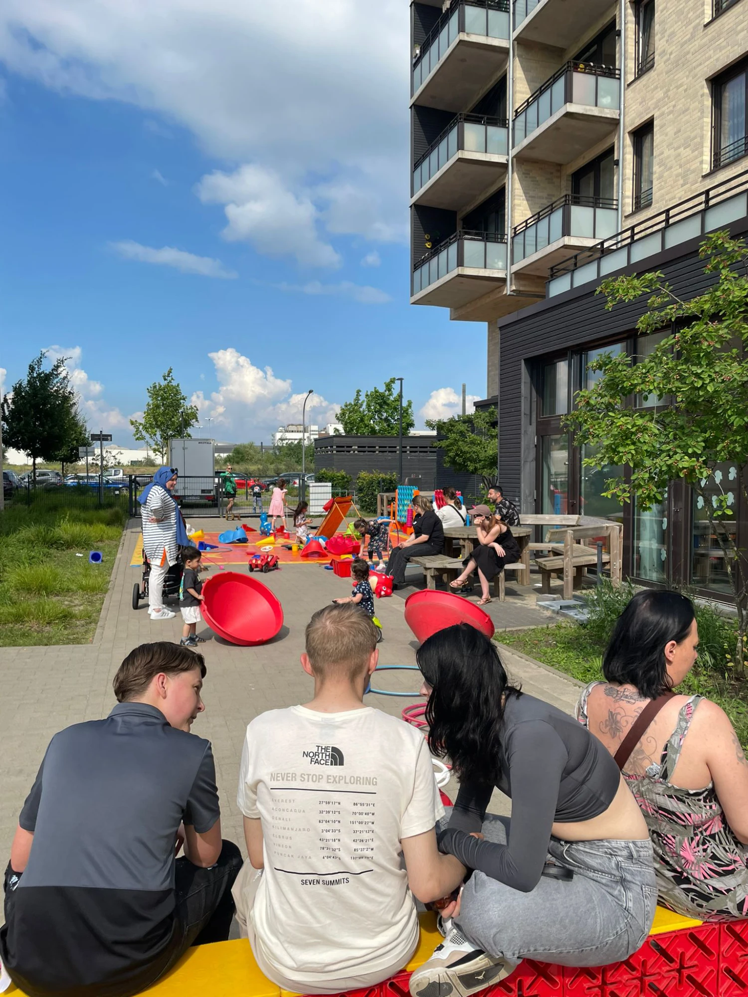 Mehrere Personen sitzen und stehen auf einem Gehweg neben einem modernen Wohngebäude mit Balkonen, im Hintergrund sind bunte Spielgeräte und Kinder zu sehen.