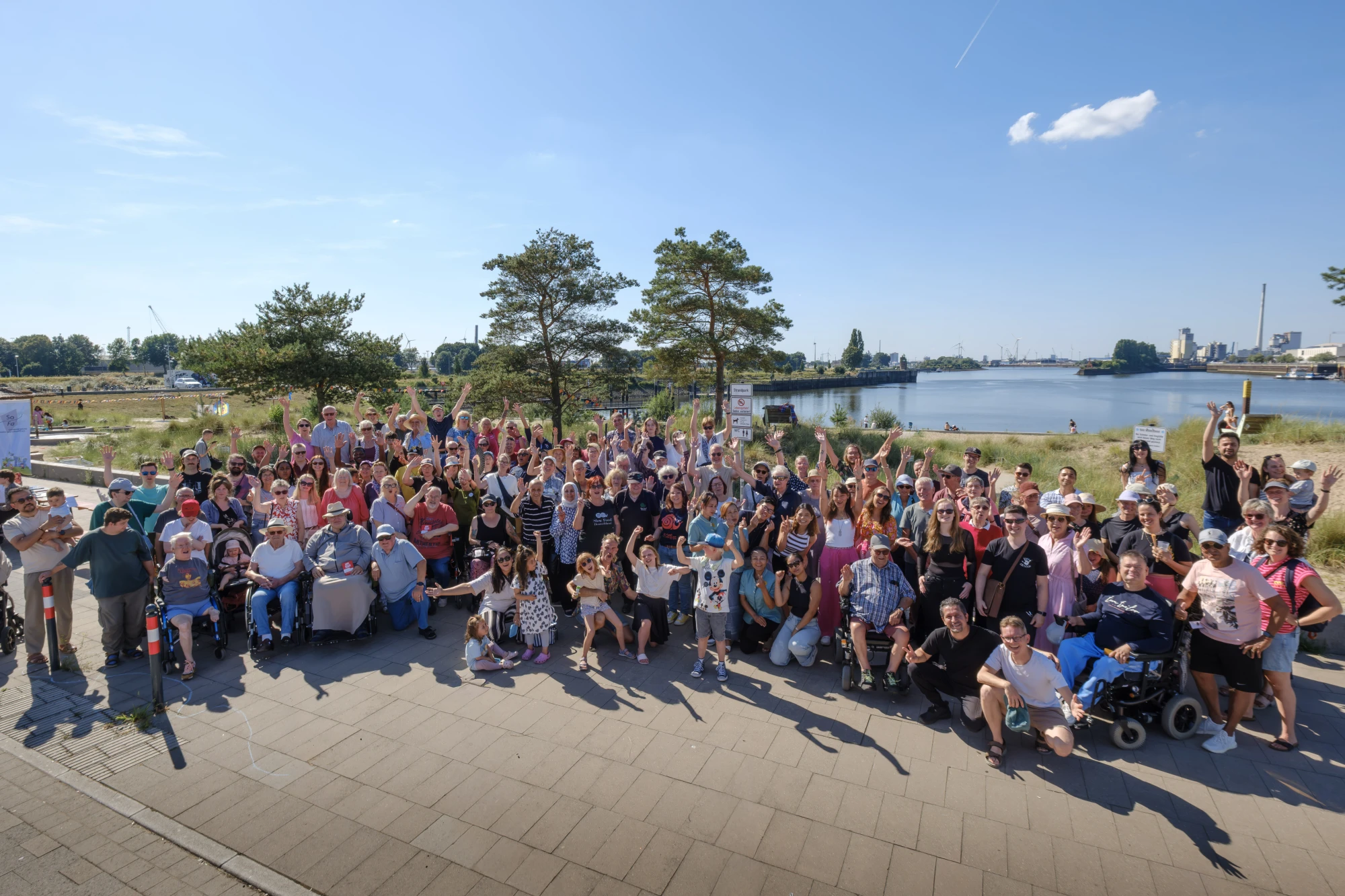 Eine gro&szlig;e Gruppe von Menschen steht und sitzt auf einem gepflasterten Platz am Wasser. Im Hintergrund wachsen B&auml;ume und Gr&auml;ser, dahinter ist ein Fluss oder See zu sehen. Viele Personen winken oder heben die Arme, einige sitzen in Rollst&uuml;hlen. Die Szene wirkt wie eine feierliche Zusammenkunft bei sonnigem Wetter. Der Himmel ist blau mit wenigen Wolken.