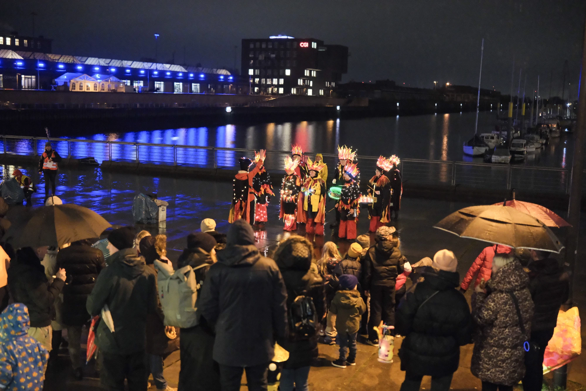 Gruppe von Menschen mit Laternen und Regenschirmen versammelt an einem Hafen, fünf Personen in bunten Kostümen stehen auf einem Steg bei Nacht.