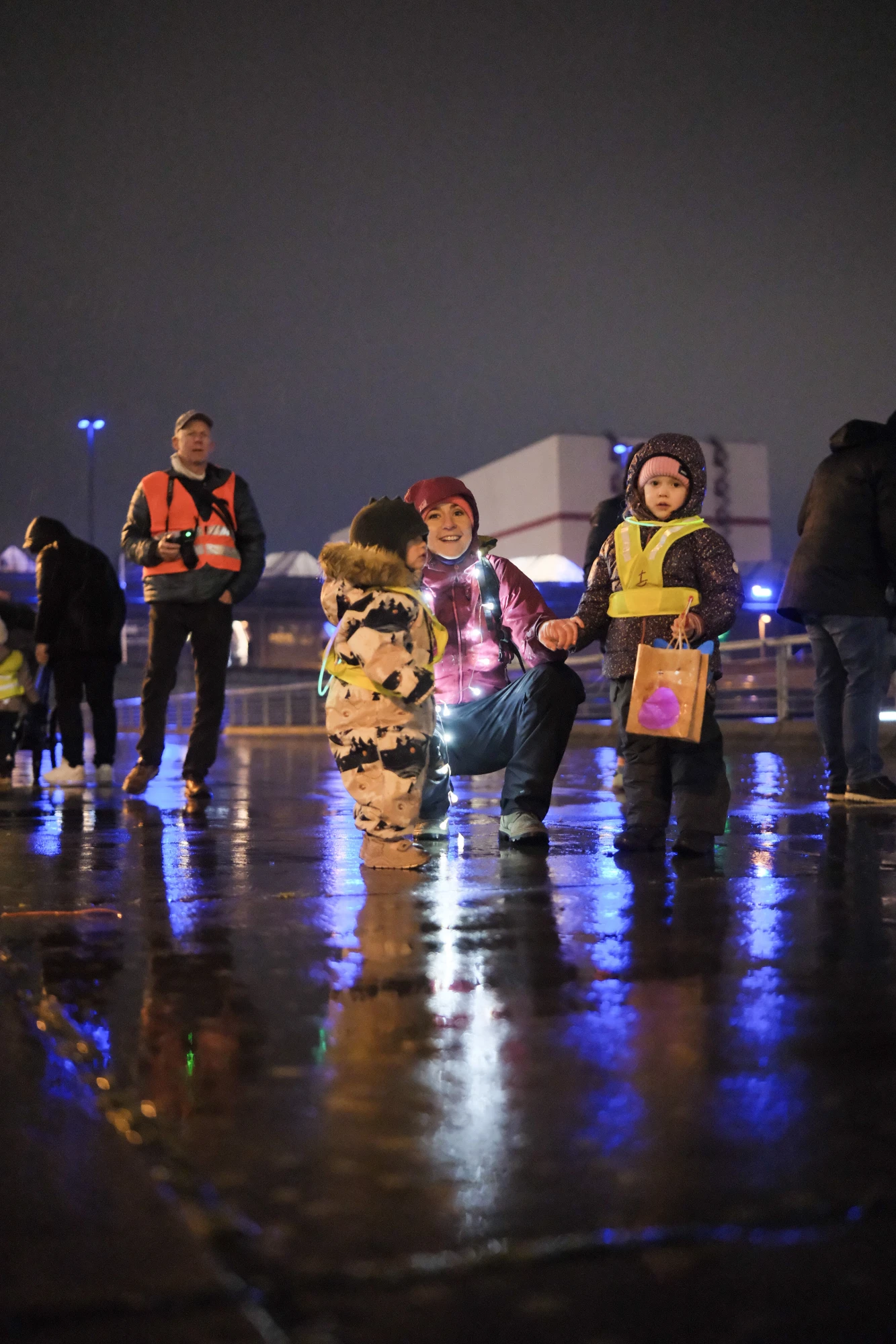 Mehrere Personen, darunter Kinder mit reflektierenden Warnwesten, stehen auf nassem Boden bei Nacht vor einem beleuchteten Gebäude.