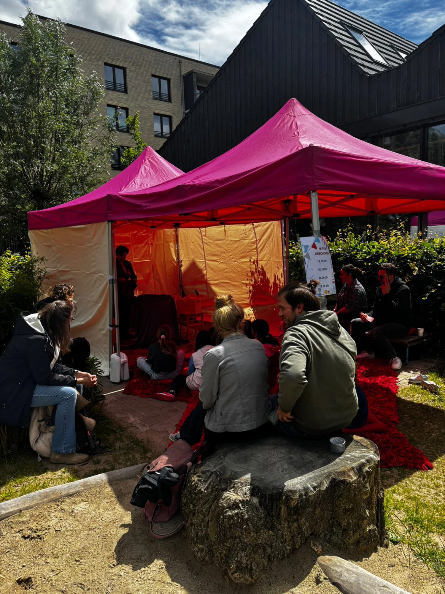 Kinder und Erwachsene sitzen und spielen unter einem pinkfarbenen Pavillon in einem Garten mit Sandkasten vor einem modernen Wohngebäude