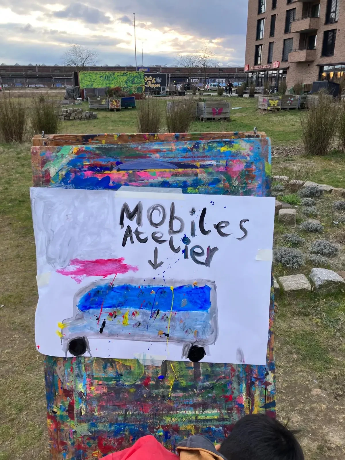 Handgemaltes Schild mit der Aufschrift 'Mobiles Atelier' und einer bunten Darstellung eines Fahrzeugs auf einer bemalten Holzfläche im Freien