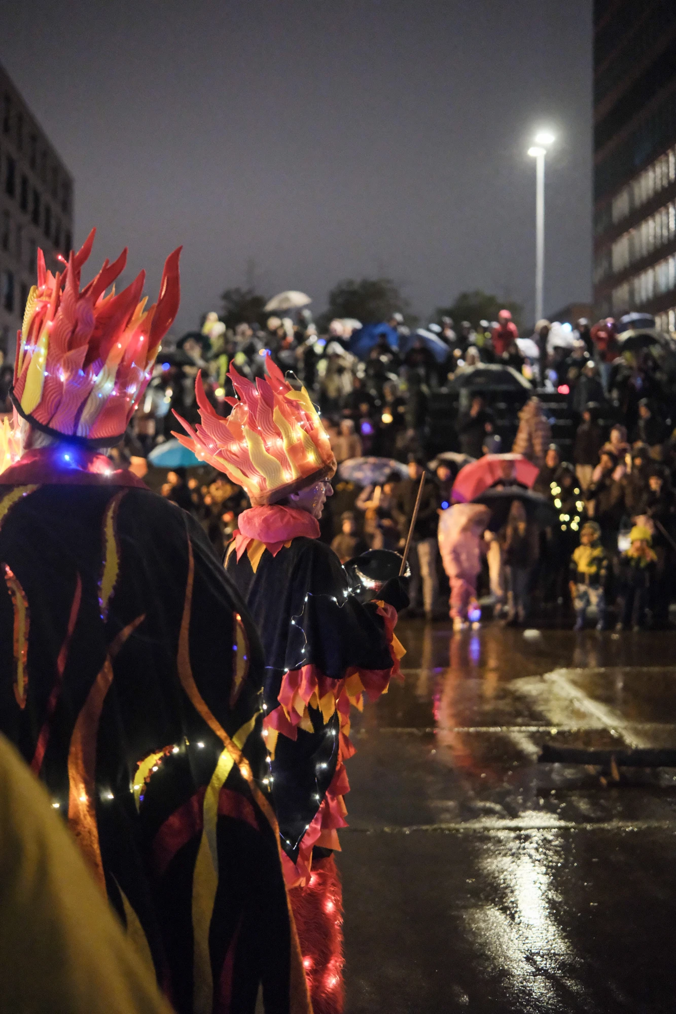 Zwei Personen in schwarzen Kostümen mit roten und orangefarbenen Flammenmuster und leuchtenden Kronen stehen vor einer Menschenmenge bei Nacht auf nasser Straße.