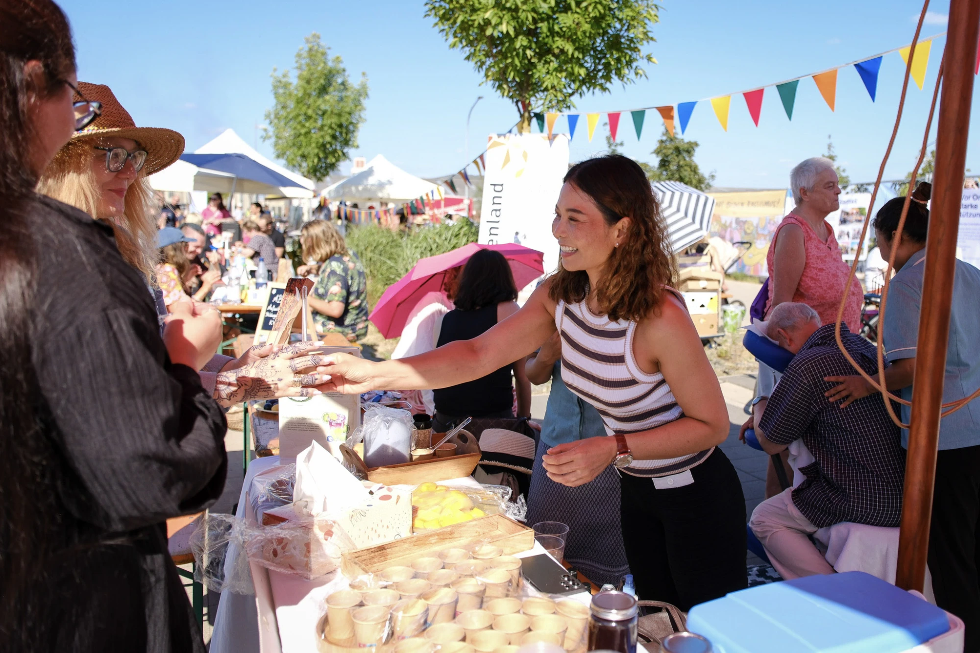 Junge Frau in gestreiftem Top reicht auf einem belebten Marktstand mit bunten Wimpeln im Hintergrund einem Kunden eine Ware
