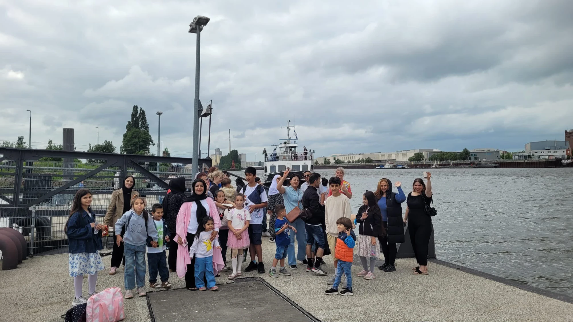 Gruppe von Menschen unterschiedlichen Alters steht auf einem Fähranleger am Wasser unter bewölktem Himmel