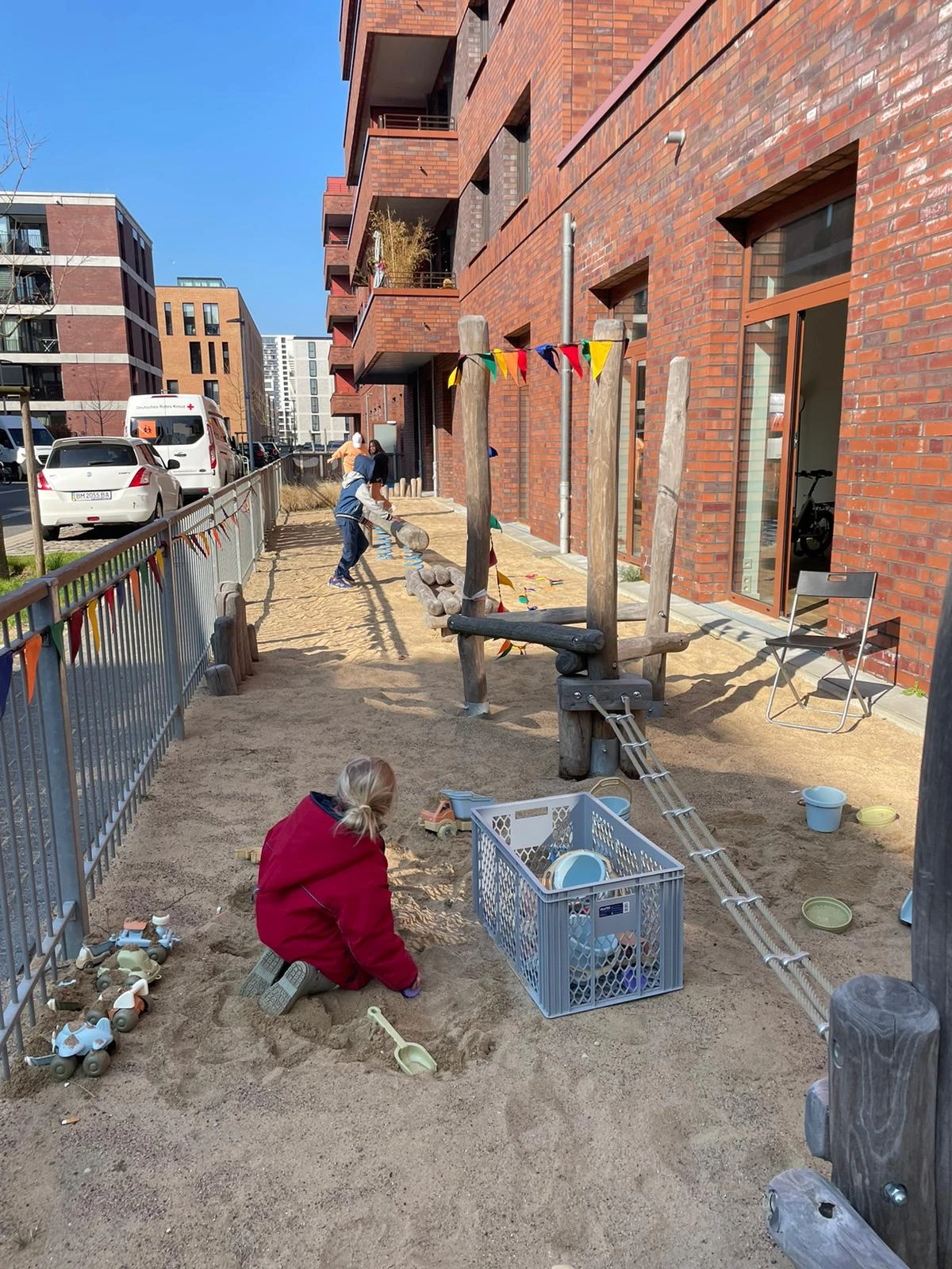 Im Sandkasten stehen verschiedene Spielger&auml;t und Sandspielzeug. Zwei Kinder spielen und toben auf dem Foto.