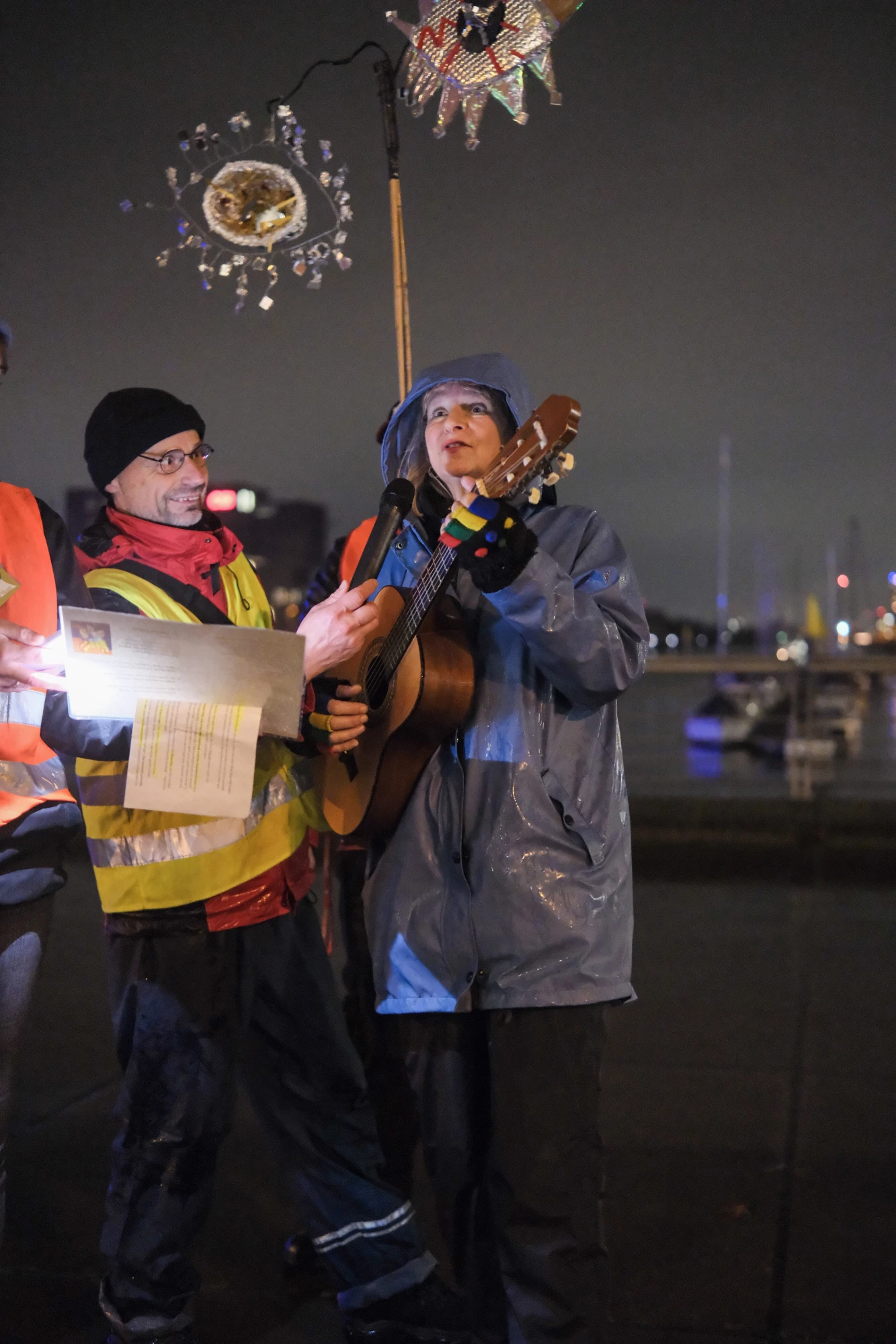 Zwei Personen bei Nacht, eine spielt Gitarre, die andere hält ein beleuchtetes Blatt Papier, im Hintergrund Laternen und Stadtlichter.