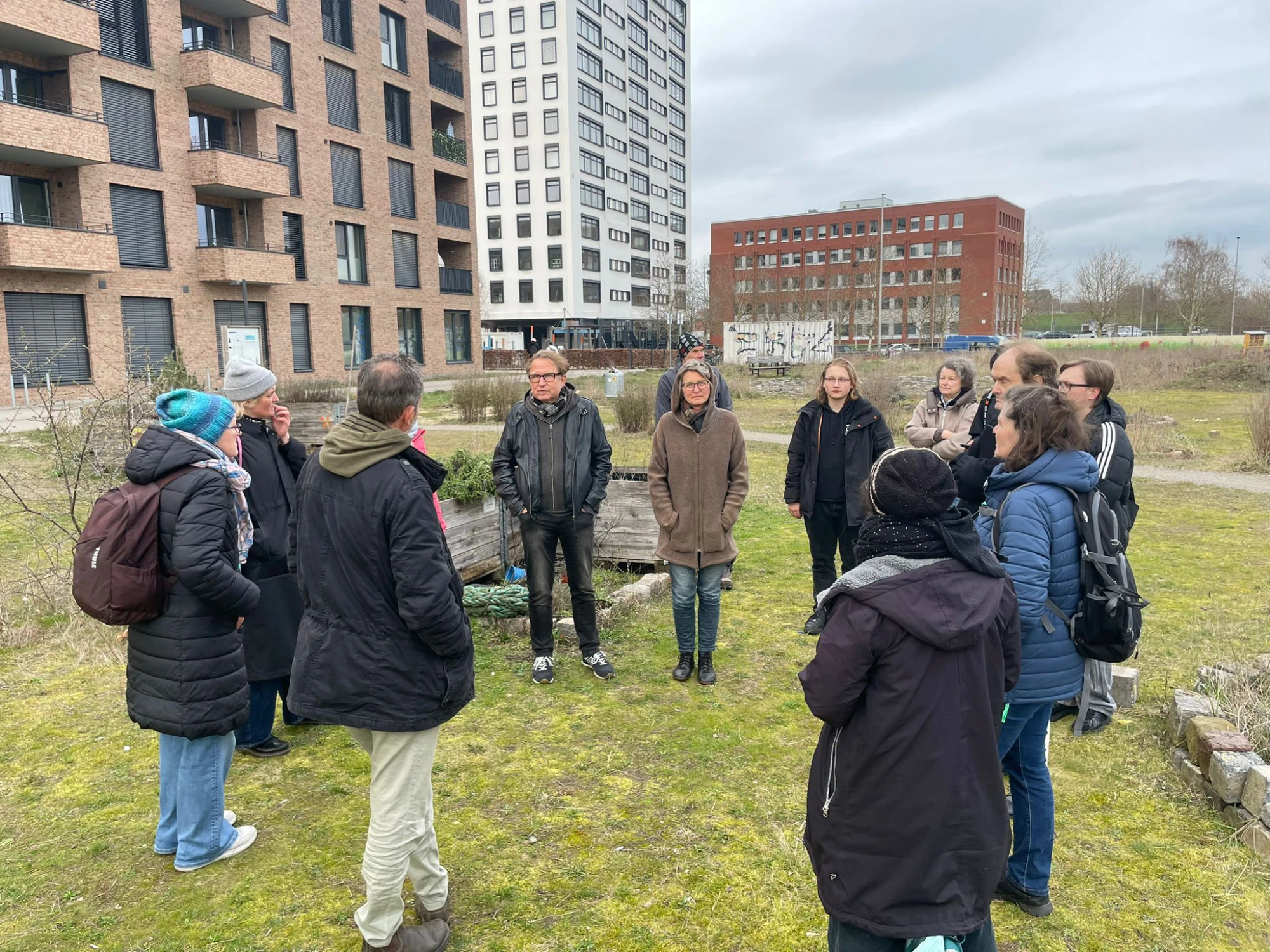 Eine Gruppe von Menschen steht im Kreis auf einer Gr&uuml;nfl&auml;che vor einem modernen mehrst&ouml;ckigen Geb&auml;ude mit dunkler Klinkerfassade. Die Personen tragen &uuml;berwiegend dunkle Winterkleidung, einige haben M&uuml;tzen oder Schals. Im Hintergrund sind weitere Geb&auml;ude mit unterschiedlichen Fassaden zu sehen. Der Himmel ist bew&ouml;lkt.