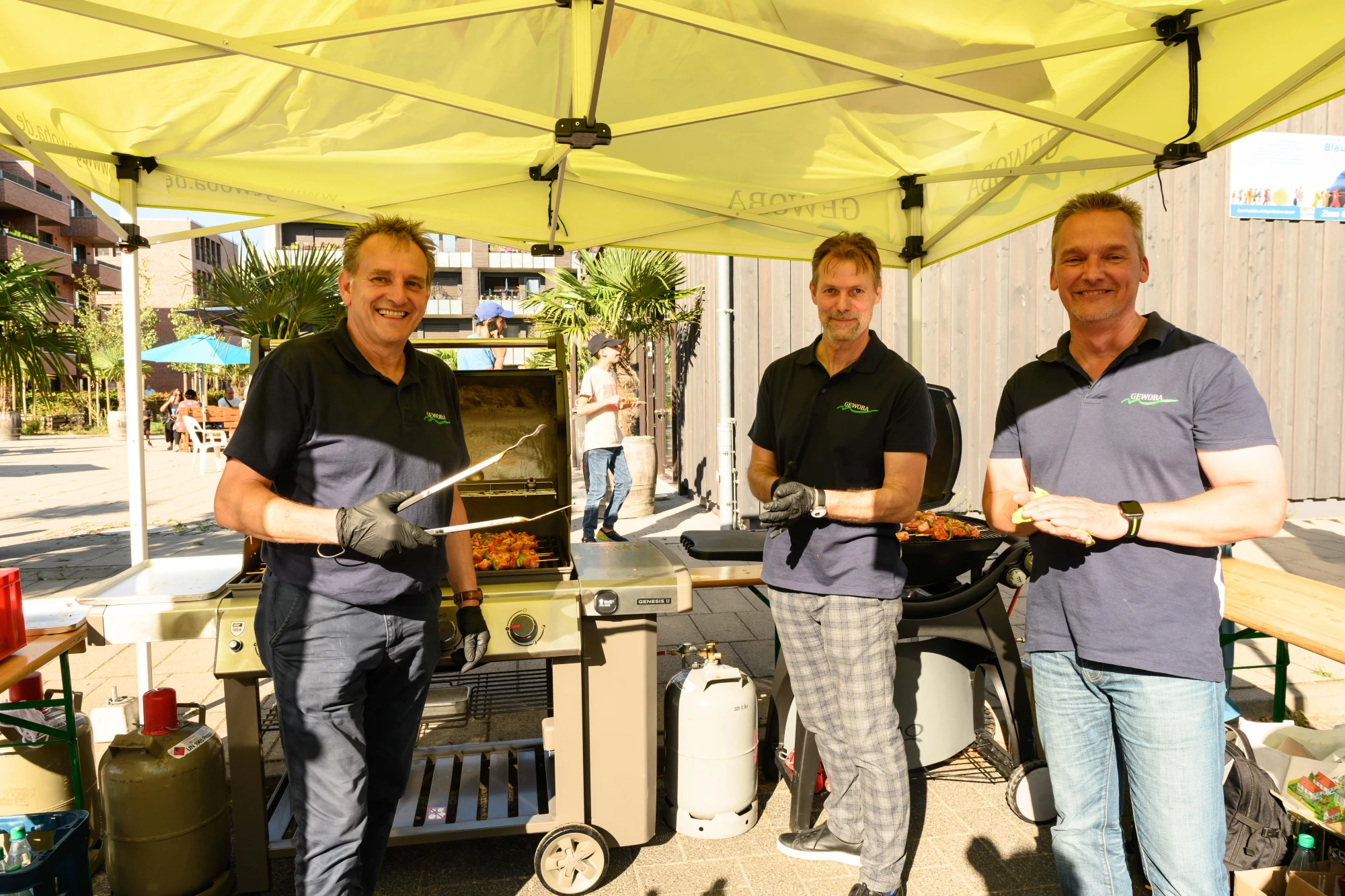 Drei Männer in schwarzen Poloshirts stehen an zwei Grills unter einem gelben Pavillon im Freien