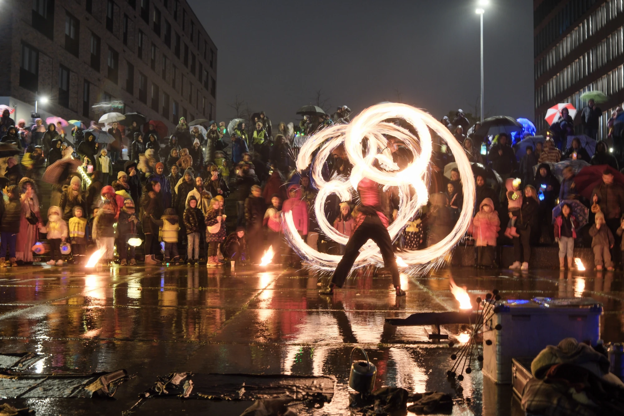 Feuerkünstler in Bewegung vor einer großen Menschenmenge bei Nacht, die mit Regenschirmen und warmen Kleidern zuschaut.