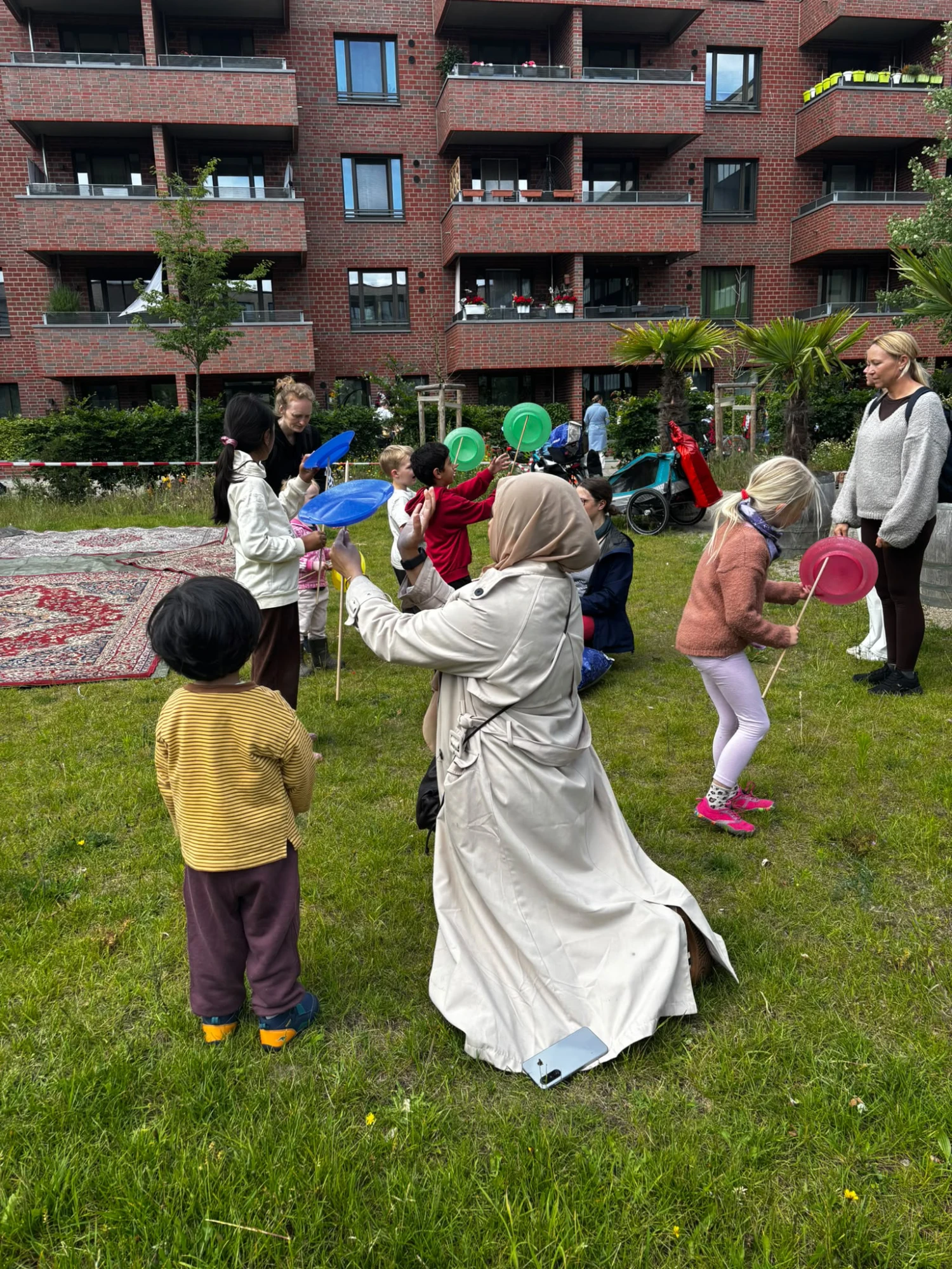 Mehrere Kinder und Erwachsene spielen auf einer Wiese vor einem mehrst&ouml;ckigen Backsteingeb&auml;ude mit bunten Tellern auf St&auml;ben