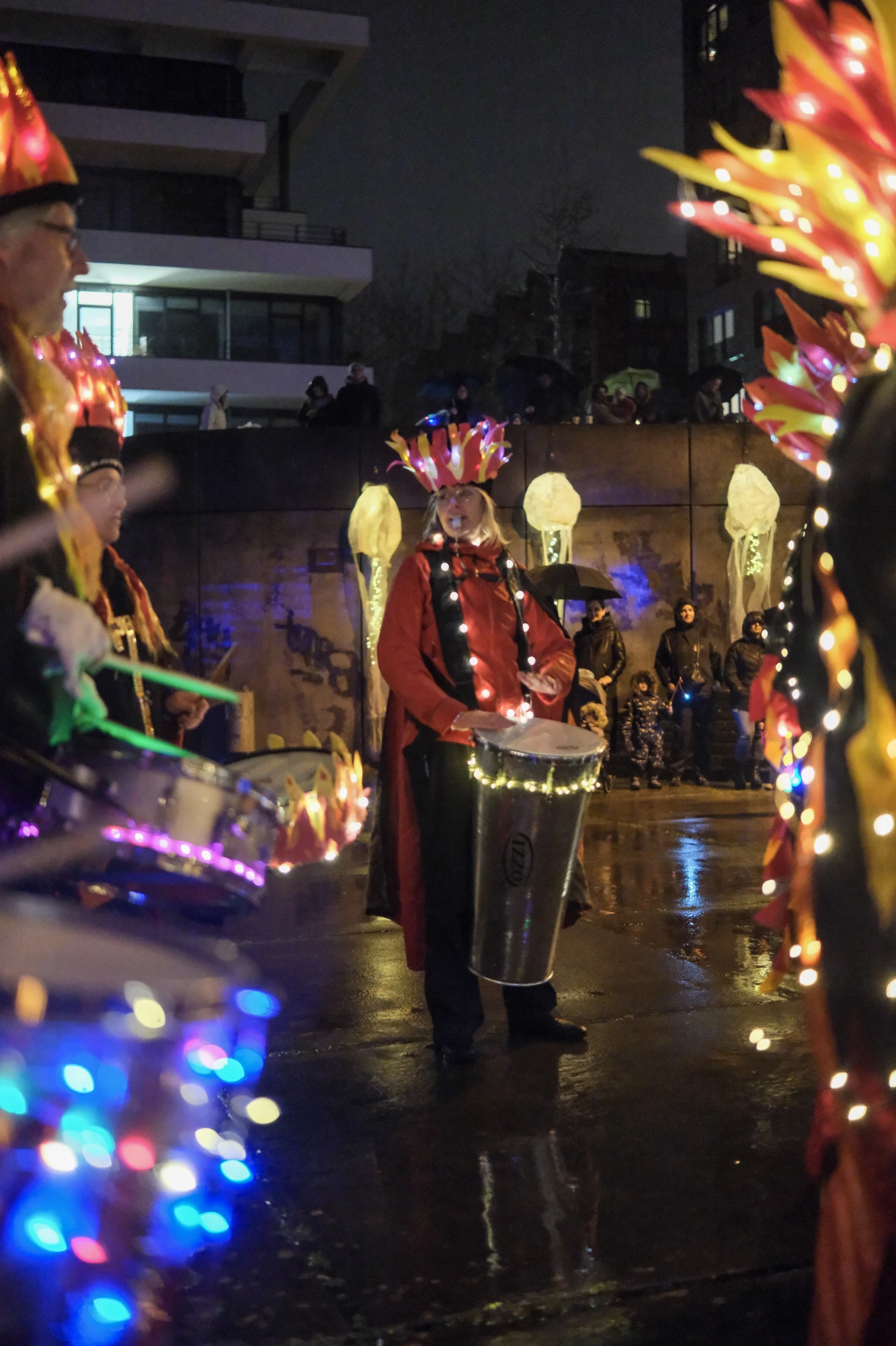 Person in rotem Kostüm mit leuchtender Kopfbedeckung spielt Trommel bei nächtlicher Veranstaltung im Freien.