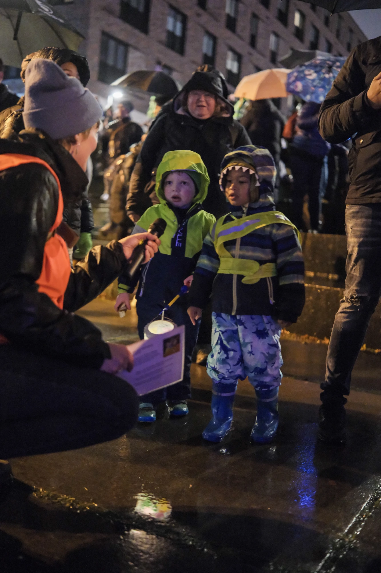 Kind in rosa Jacke und Gummistiefeln steht auf nassem Boden, umgeben von mehreren Personen, eine hält einen Regenschirm.