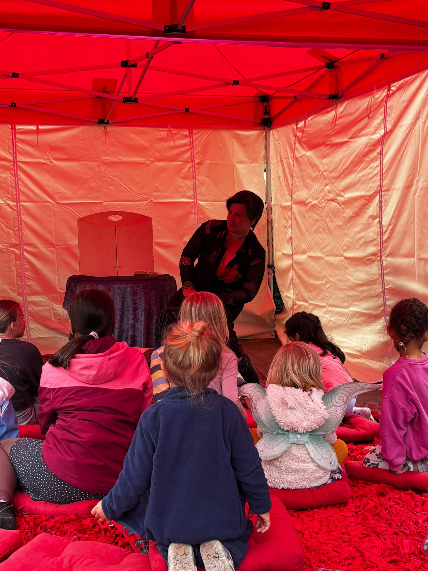 Kinder sitzen auf roten Kissen in einem Zelt und hören einer vorlesenden Person zu