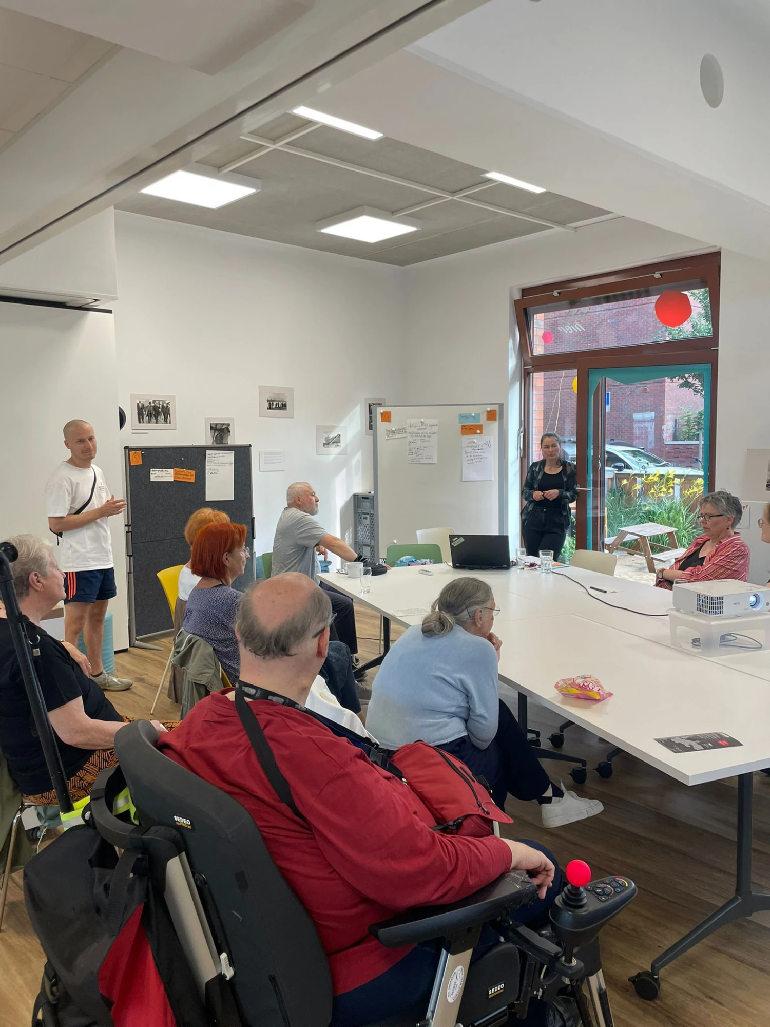 Zu sehen sind 10 Personen in einem Raum, die rund um einen Tisch sitzen bzw. stehen und sich austauschen. Im Hintergrund sind 2 Pinnw&auml;nde zu sehen, an denen Zettel gepinnt sind.