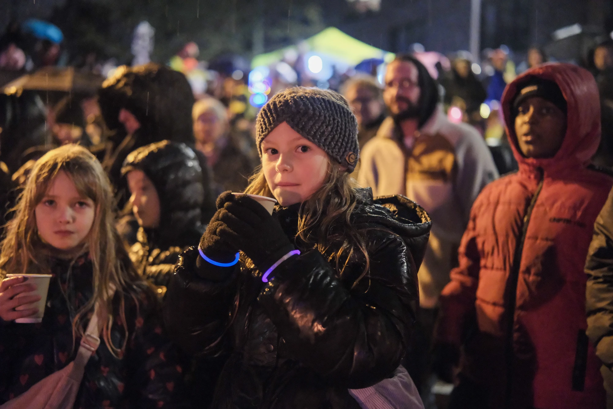 Gruppe von Menschen bei Nacht, einige tragen Winterjacken und Mützen, im Hintergrund bunte Lichter und ein gelbes Zelt.