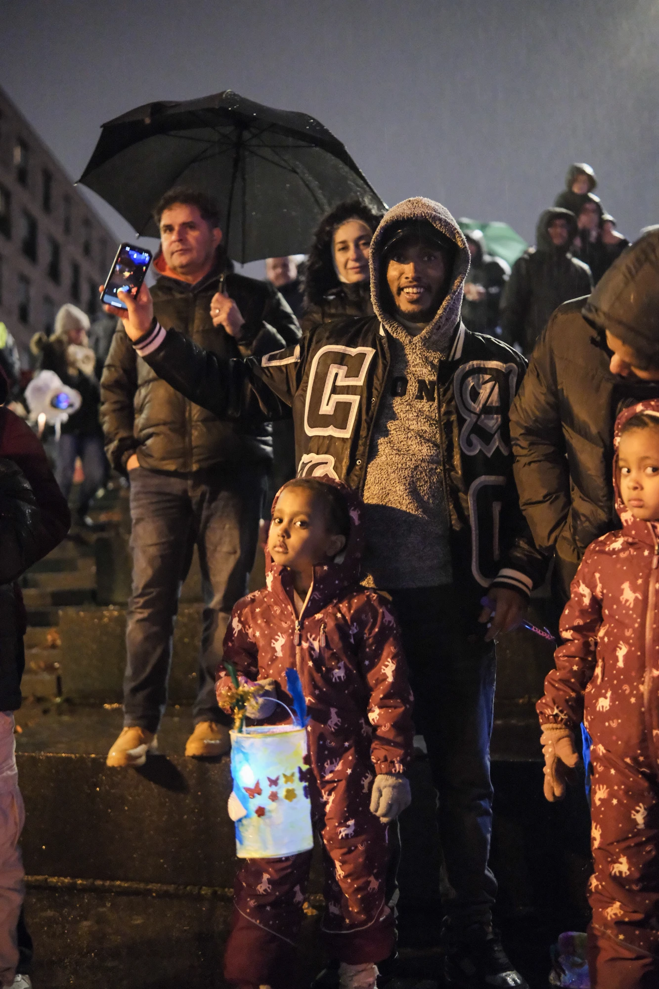 Mehrere Personen bei Nacht, darunter zwei Kinder in roten Anzügen mit Laternen, eine Person hält einen schwarzen Regenschirm und ein Smartphone hoch.