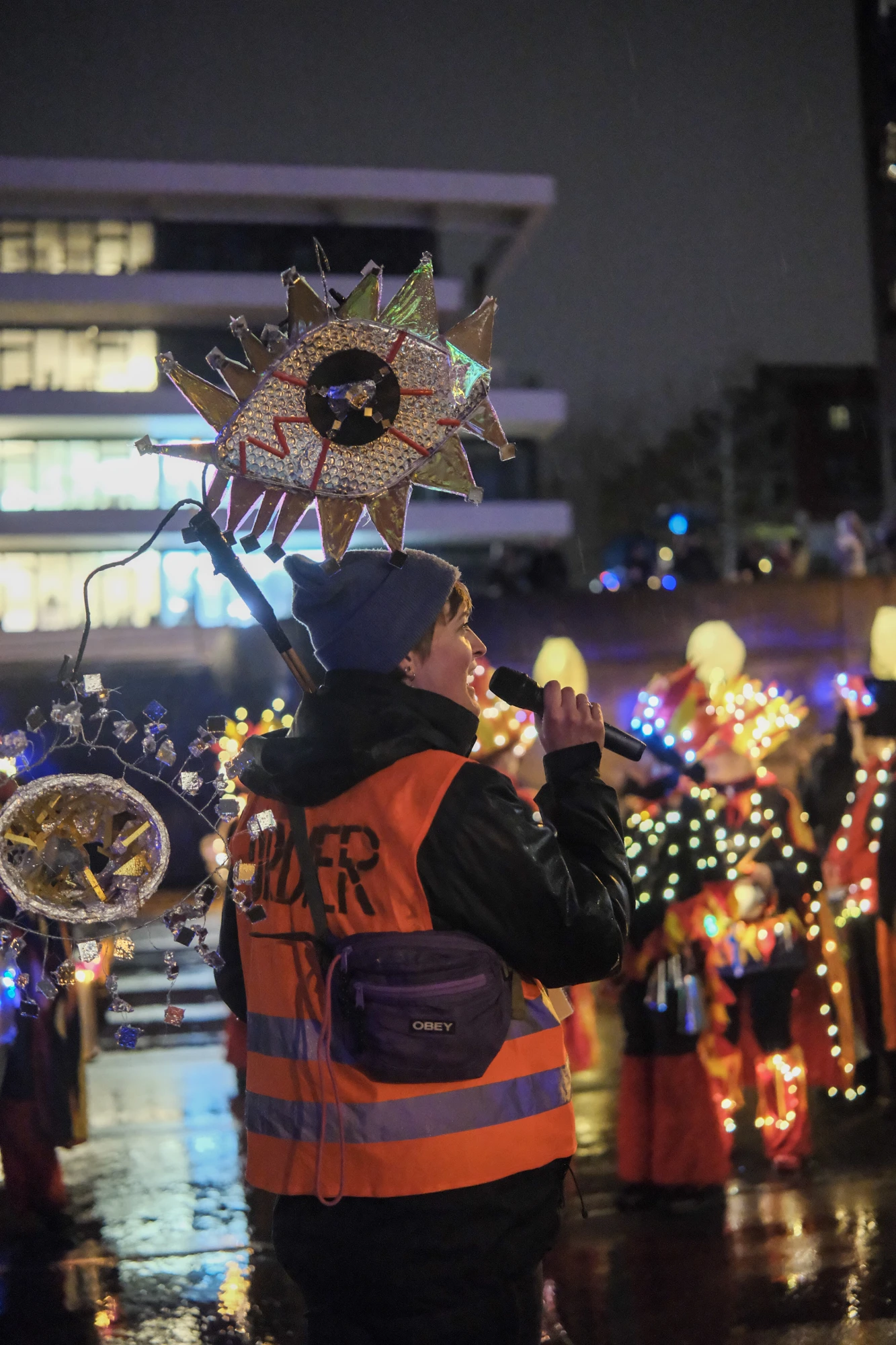 Person mit orangefarbener Warnweste hält eine Laterne in Form eines stilisierten Auges mit bunten Zacken bei Nacht.