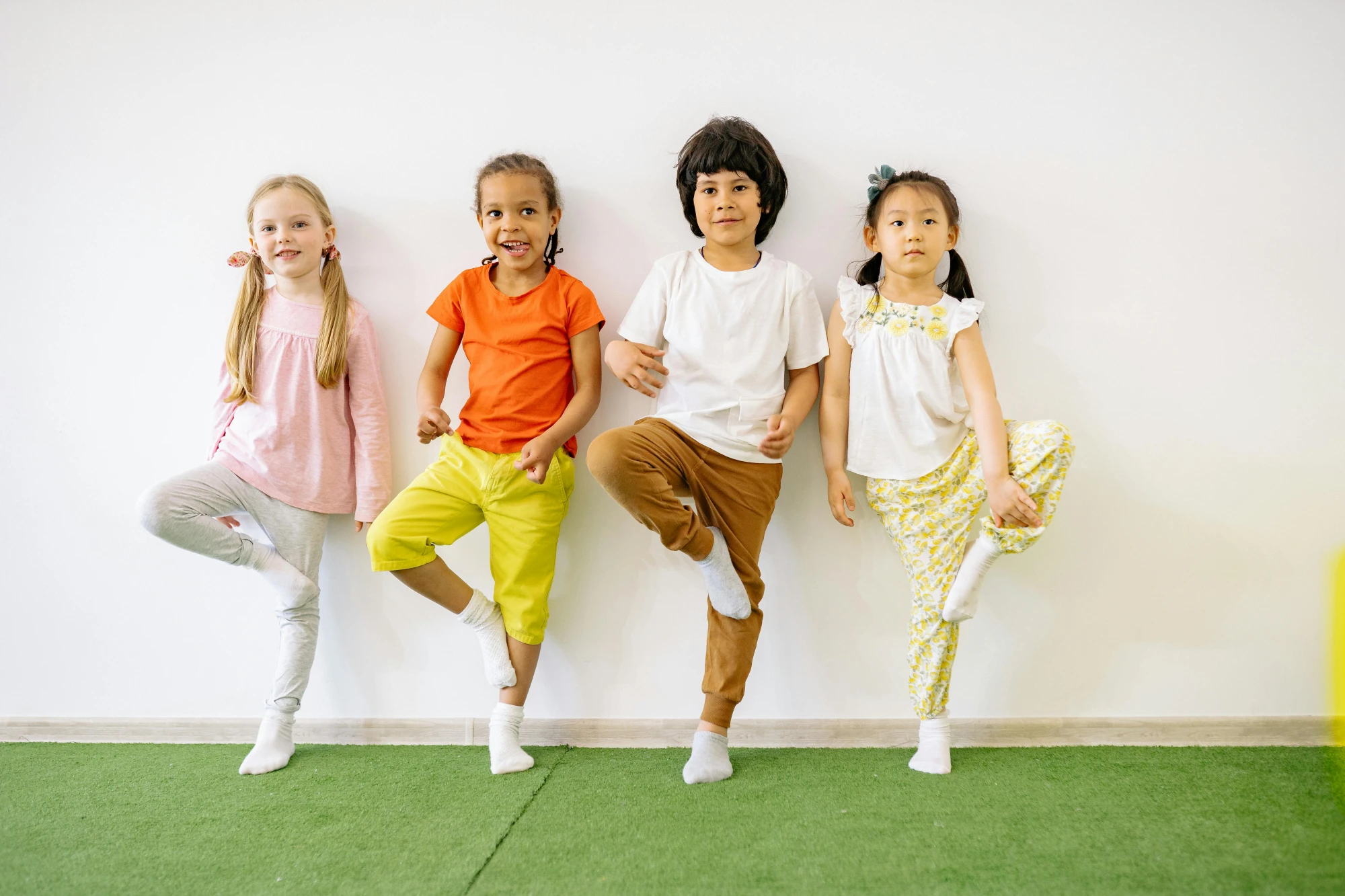 Vier Kinder stehen nebeneinander auf grünem Teppich und balancieren jeweils auf einem Bein mit erhobenem anderen Bein
