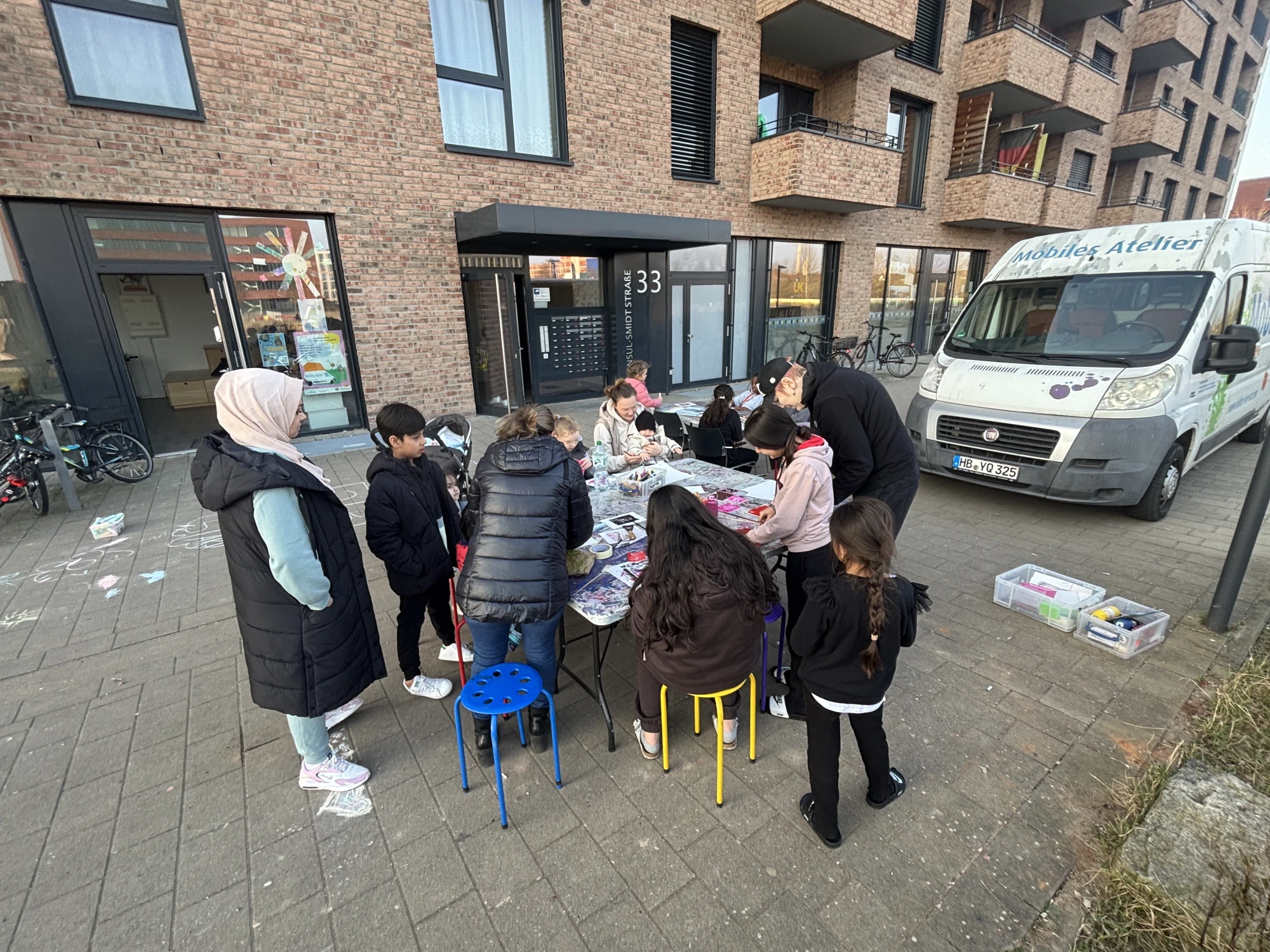 Das Mobile Atelier steht vor einem Haus. Davor sitzen Kinder und Erwachsene an einem Tisch und basteln.