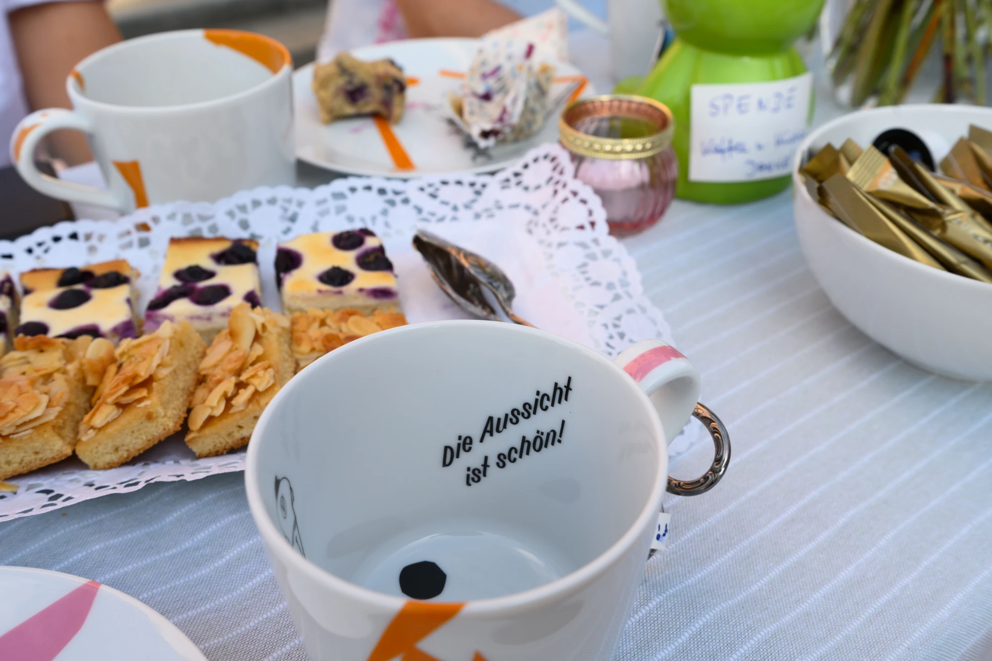 Tasse mit der Aufschrift 'Die Aussicht ist schön' auf einem gedeckten Tisch mit Gebäck, Tasse und Schale mit goldfarbenen Stäbchen