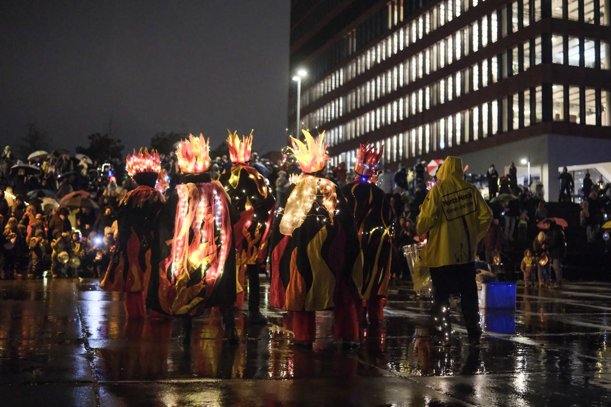 Gruppe von sechs Personen in leuchtenden Kostümen mit Flammenmotiven, eine Person spielt Trommel, nächtliche Stadtkulisse im Hintergrund.