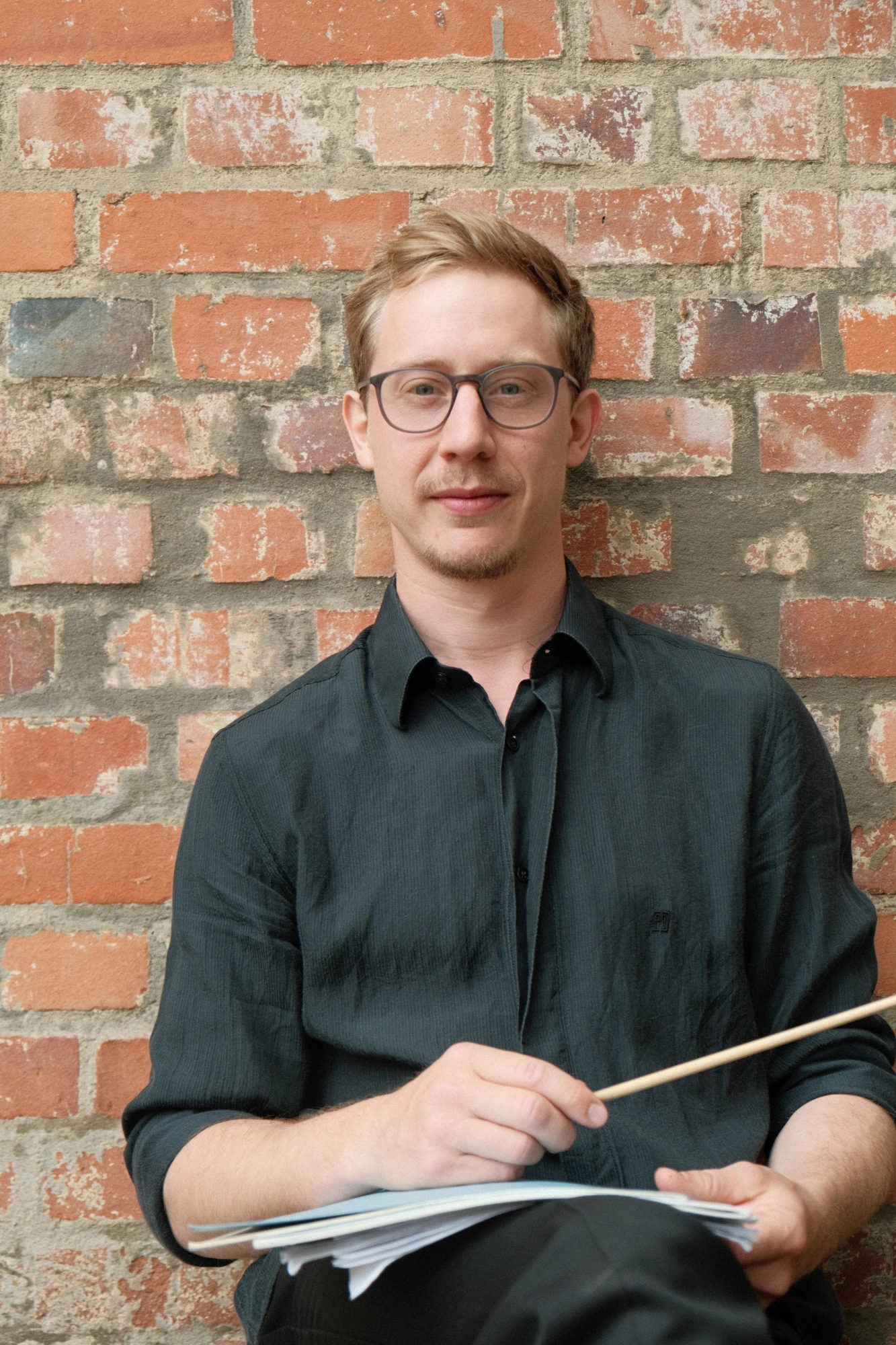 Ein junger Mann sitzt vor einer Backsteinwand, hält einen Dirigierstab in der Hand und schaut entspannt in die Kamera.